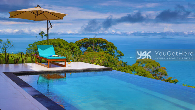 View of the infinity pool and ocean with lounge chair under umbrella on the pool deck.
